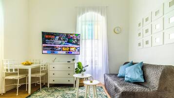 Uma sala de estar com televisão de tela plana exibindo conteúdo da Netflix, uma mesa de jantar branca com cadeiras, uma pequena mesa redonda com uma planta, um sofá cinza com almofadas azuis e um tapete estampado.