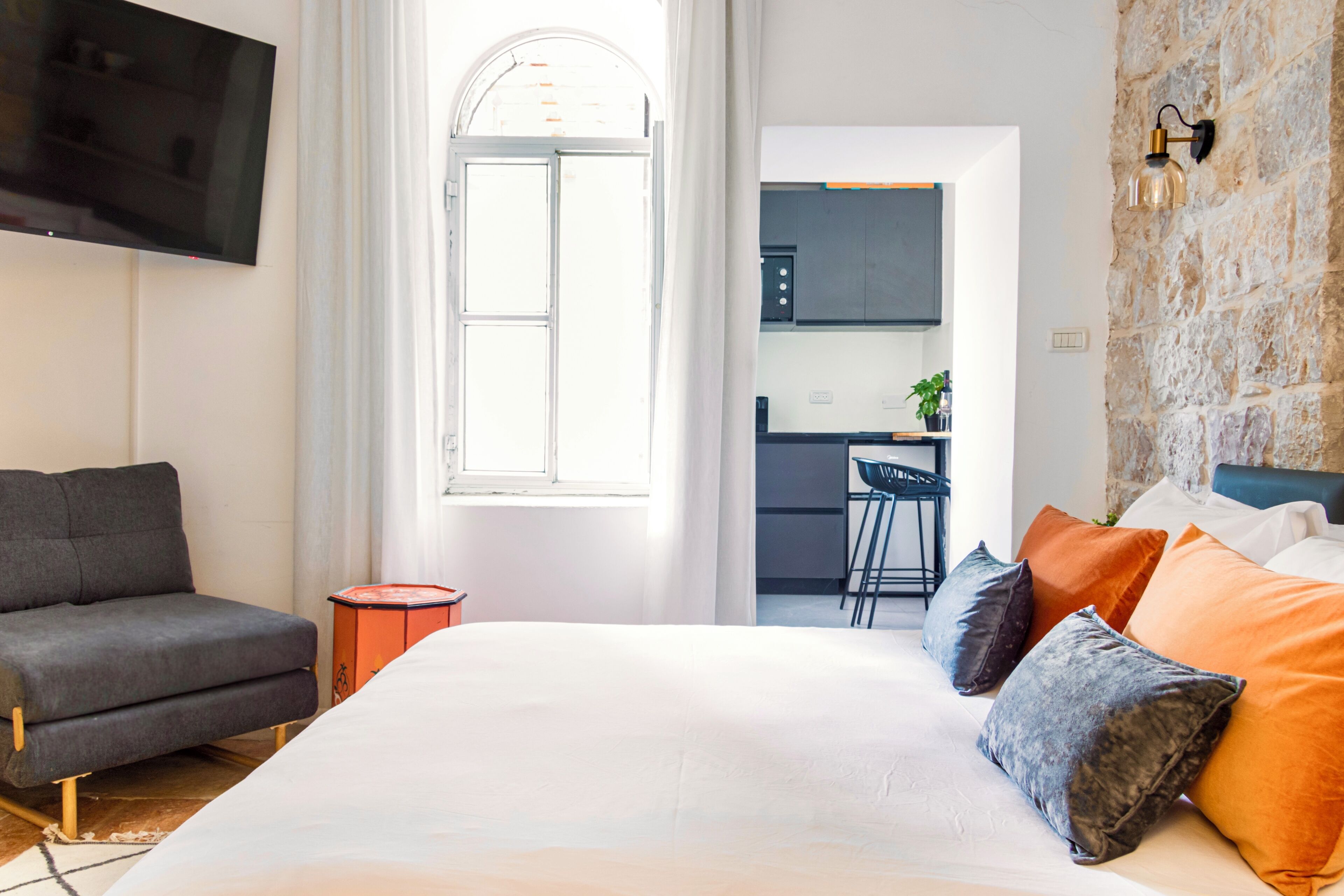 A modern hotel room with a large bed, a grey sofa, a TV, a kitchen area, and a stone wall.