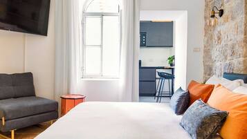 A modern hotel room with a large bed, a grey sofa, a TV, a kitchen area, and a stone wall.