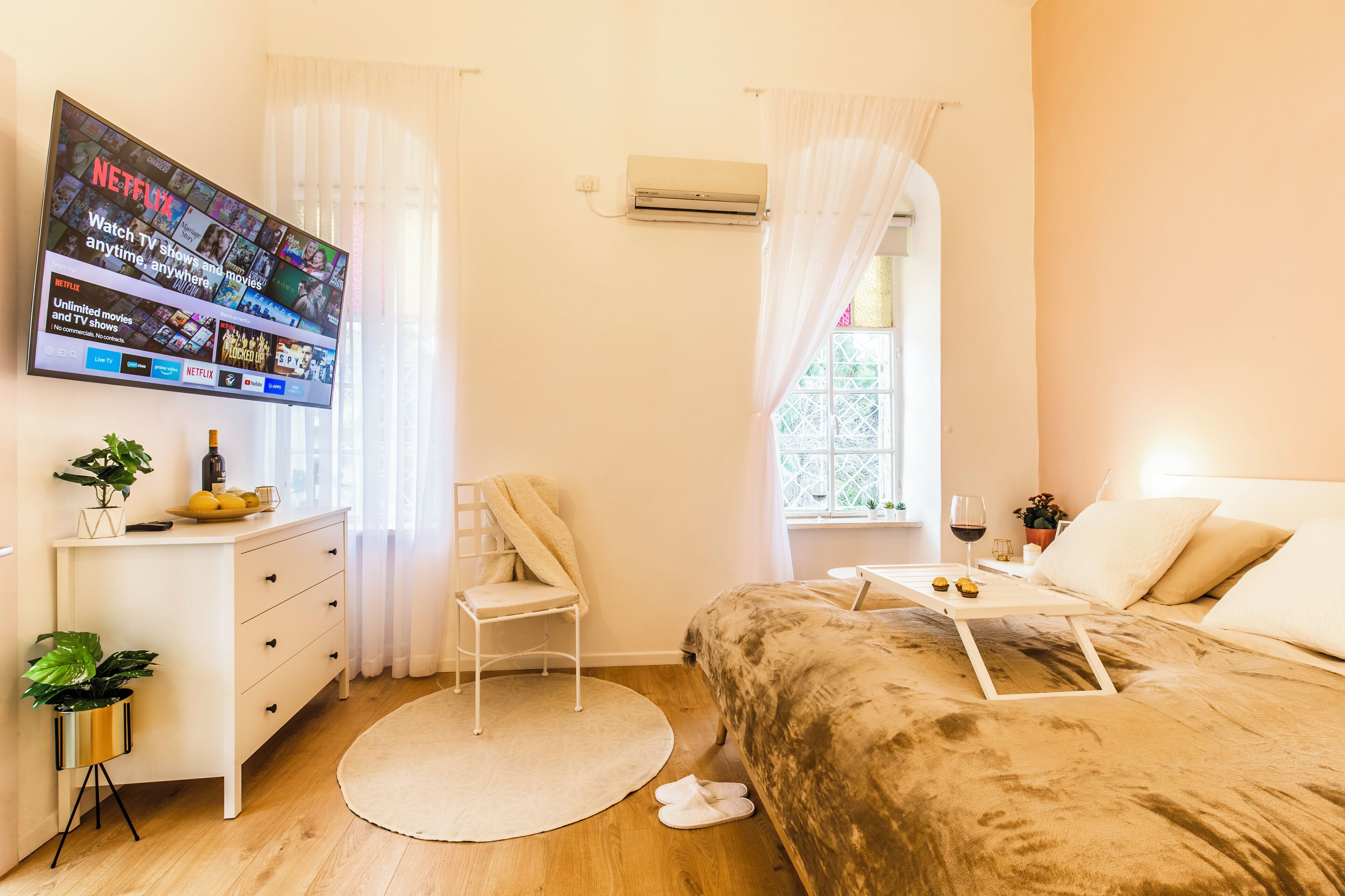 A bedroom with a bed, a TV mounted on the wall, a desk with a chair, and a window with curtains.