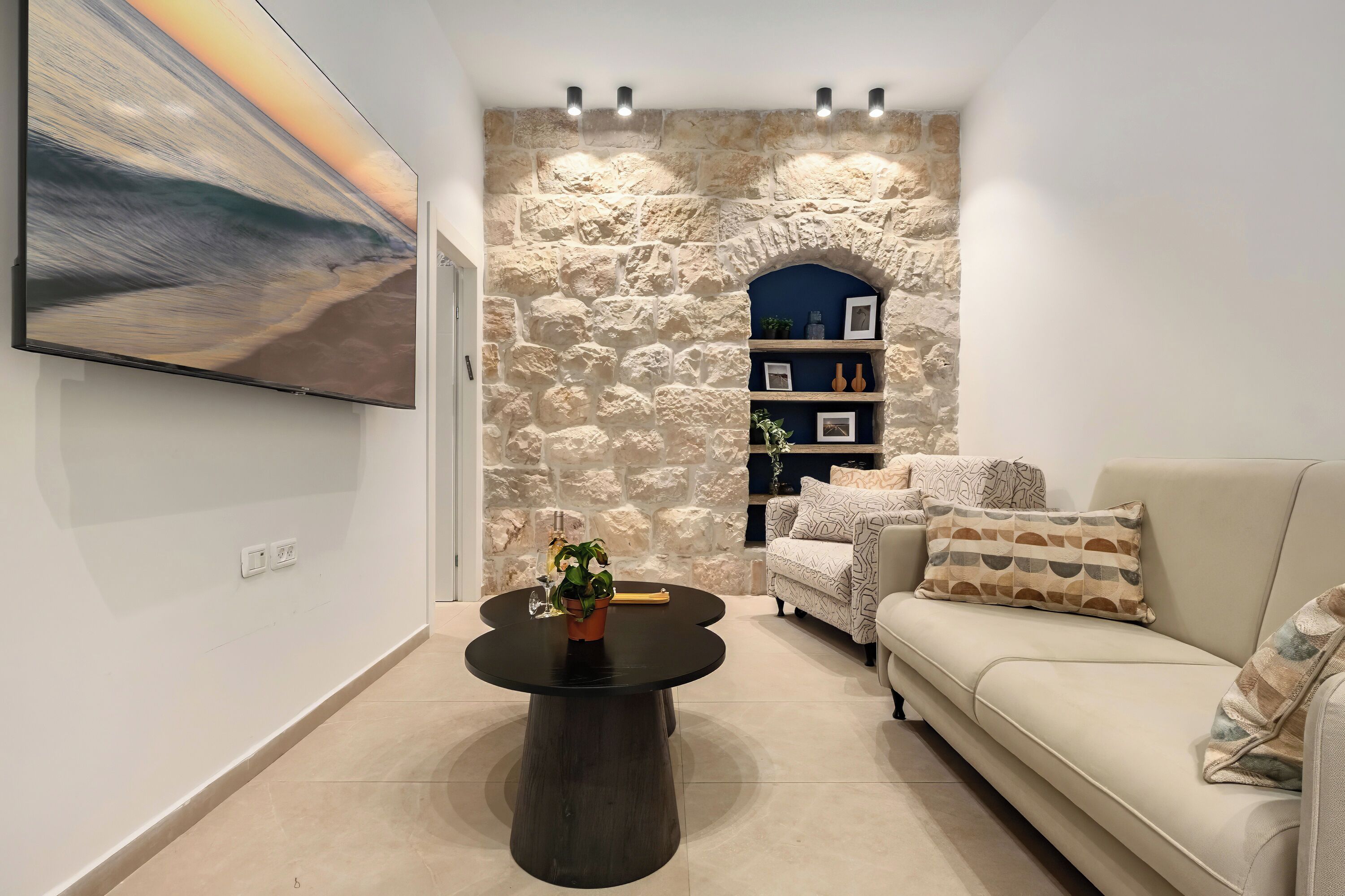 A modern living room with a stone wall, a flat-screen TV, a round coffee table with a plant, a beige sofa, and a bookshelf.