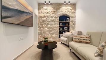 A modern living room with a stone wall, a flat-screen TV, a round coffee table with a plant, a beige sofa, and a bookshelf.