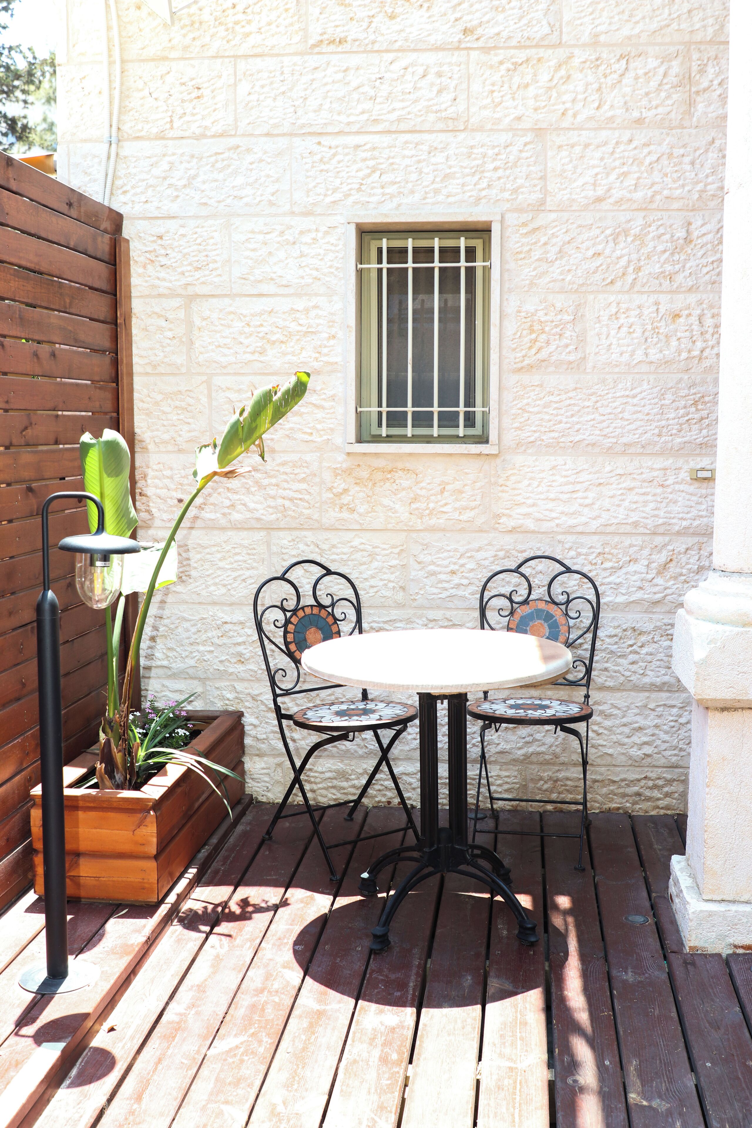 Un patio al aire libre con una mesa de madera y sillas de metal, una planta en maceta y una ventana con barrotes.