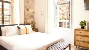 Une chambre avec un grand lit, un banc en bois, une commode en osier et une vue sur le paysage, visible à travers une fenêtre en arc de cercle.