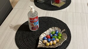 Un tavolo da pranzo con sottopiatti neri, una bottiglia di disinfettante per le mani, un bicchiere trasparente con una rosa rossa e un piatto di cioccolatini confezionati.