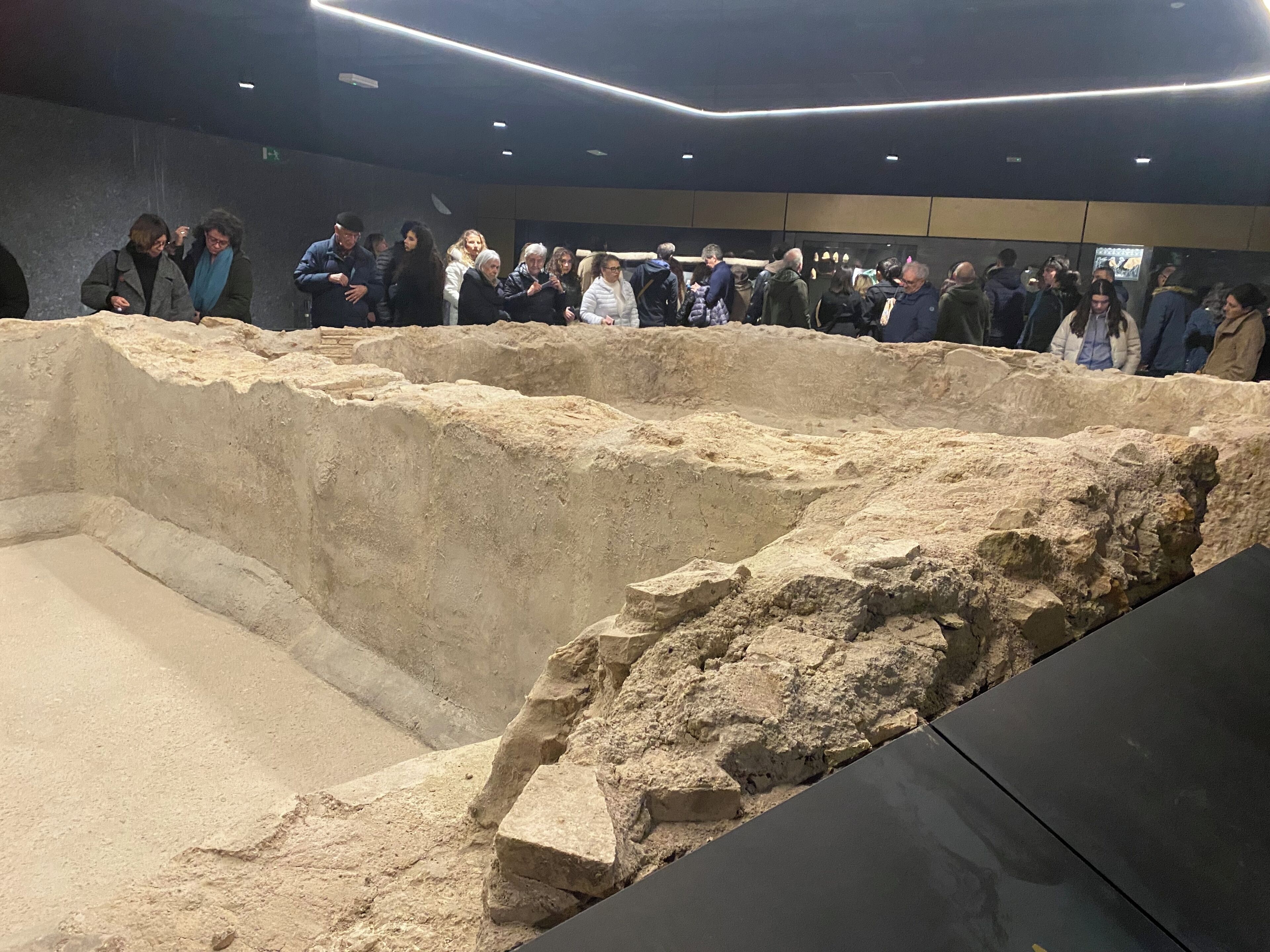 Un espai interior d'una exposició arqueològica amb visitants observant una gran excavació.
