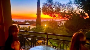 Un balcon offrant une vue sur un coucher de soleil se reflétant sur une étendue d’eau, avec un grand arbre et un bâtiment en arrière-plan.