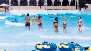 Une piscine où l’on voit des personnes dans l’eau et au bord, entourée d’un bâtiment doté de fenêtres en arc de cercle.