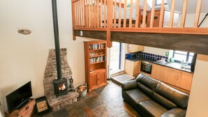 A cozy living room with a stone fireplace, a wooden bookshelf, a leather sofa, and a kitchen area with a window. - The Hay Barn (High Peak)