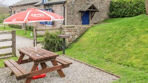 A stone house with a wooden door and windows, a picnic table with a red umbrella, and a grassy hill in the background. - The Hay Barn (High Peak)