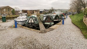A canal-side boat moored in a gravel dock area with a stone building in the background. - The Hay Barn (High Peak)