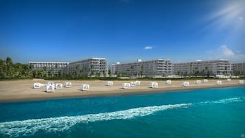 A beachfront hotel with multiple buildings, a sandy beach with white sunbeds, and a clear blue ocean.