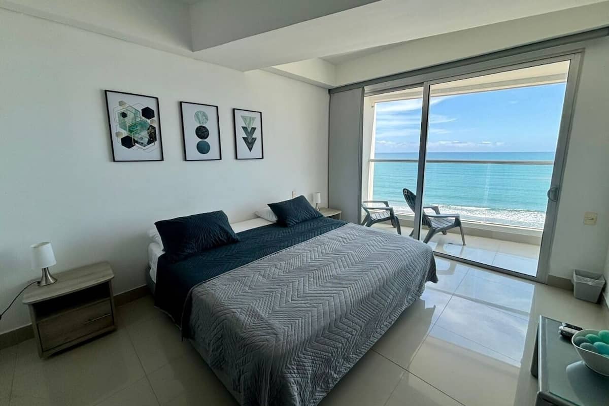 A modern bedroom with a large bed, a nightstand, a desk, and a balcony with a view of the ocean.