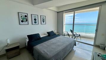A modern bedroom with a large bed, a nightstand, a desk, and a balcony with a view of the ocean.