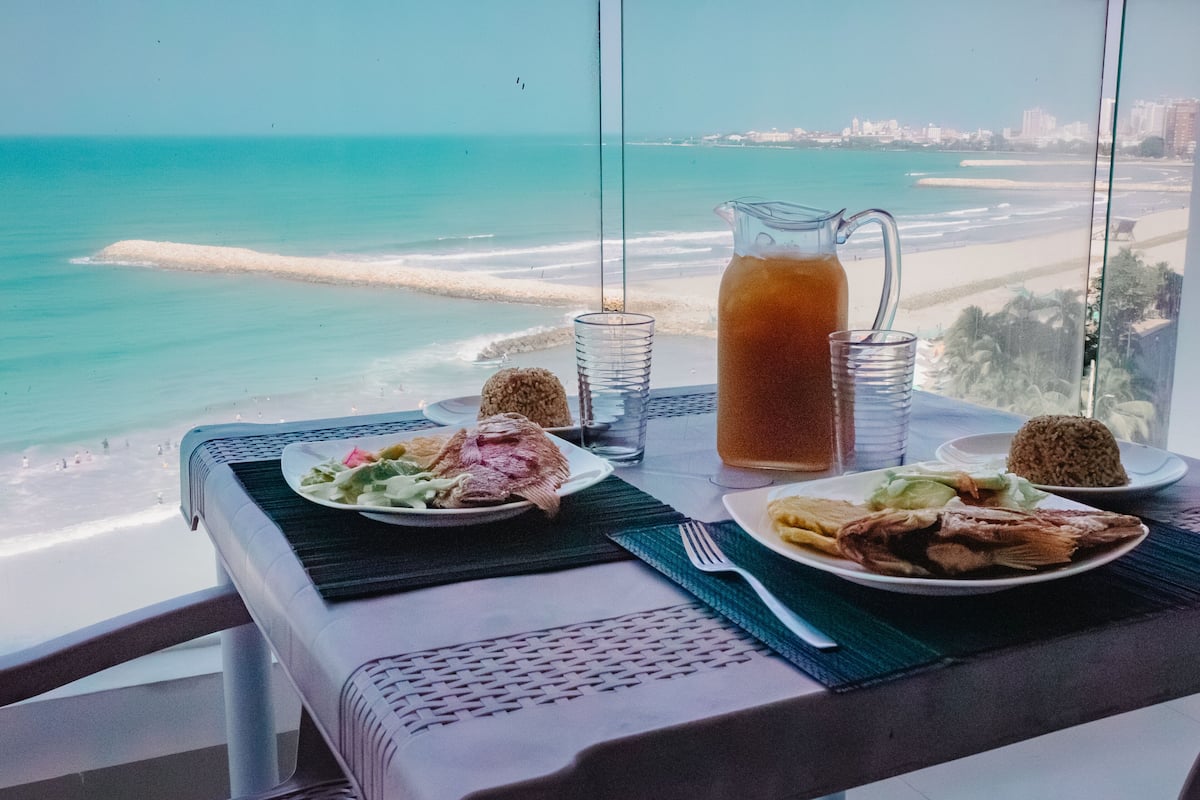 A table with plates of food, a pitcher of iced tea, and a glass of water, overlooking a beach and ocean view.