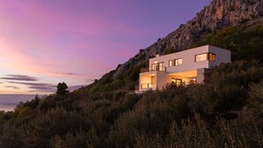Una casa moderna de dos pisos con balcón, rodeada de vegetación densa y un acantilado rocoso.