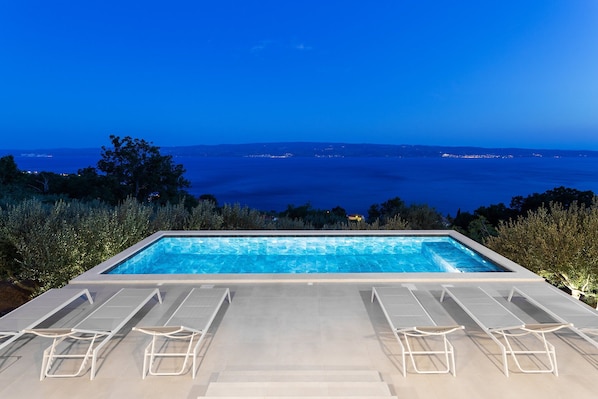 A rectangular infinity pool with clear blue water, surrounded by white lounge chairs, set against a backdrop of a mountainous landscape and a view of the sea at dusk.