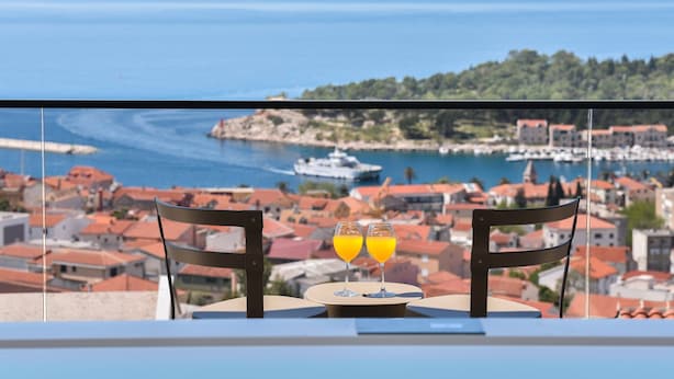 A table with two glasses of orange juice, a chair, and a view of a coastal town with buildings and a marina. - Luxury Villa Persephone with Pool (Makarska)