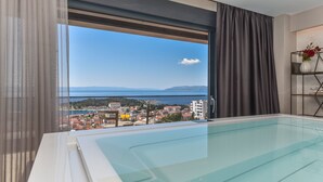 A modern hotel room with a large window, a glass-enclosed pool, and a view of a coastal town. - Luxury Villa Persephone with Pool (Makarska)