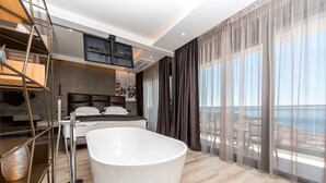 Un dormitorio moderno con una bañera grande, una cama con cabecero y vistas al mar a través de cortinas translúcidas.