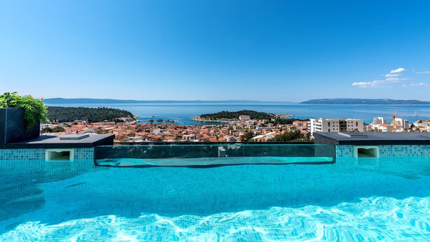 Una piscina con agua cristalina azul, rodeada por un paisaje urbano y vistas al mar.