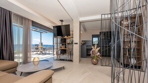 Una sala de estar moderna con un sofá, una mesa redonda de centro y un televisor. Hay un balcón con vista al mar.