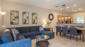 A modern living room with a sectional sofa, a coffee table, and a dining area with chairs and a table. There are abstract paintings on the wall and a kitchen in the background. - Stylish 3BR 2.5BA Townhome Free Tub not overlooked (Kissimmee)