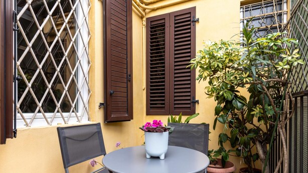 A small outdoor seating area with a round table and chairs, a potted plant with flowers, and a window with a lattice design. - Appartamento Alla Finestrella by Wonderful Italy (Bologna)