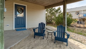 A covered porch with two Adirondack chairs and a small table, a blue door with a wreath, and a gravel driveway. - 🌟Charming Retreat🌟 Dogs ✔️, Beach Chairs, Bikes, Bonus Living Room, ❤️ of KDH (Kill Devil Hills)