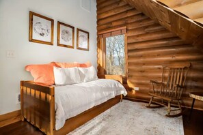 Quarto de cabana de toras com uma cama, uma cadeira de balanço e três quadros na parede.