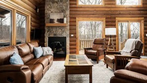L'intérieur d'une cabane en rondins, avec une cheminée en pierre, des canapés en cuir, une table basse et une grande fenêtre donnant sur une forêt.