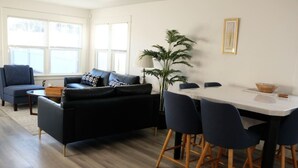 A modern living room with a black sofa, a dining table with four chairs, and a large window with blinds. - Aviation Landing Vacation Homes Fallbrook CA (Fallbrook)