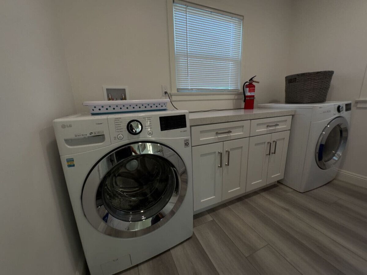 A laundry room with two front-loading washing machines, a dryer, a window with blinds, and a fire extinguisher.