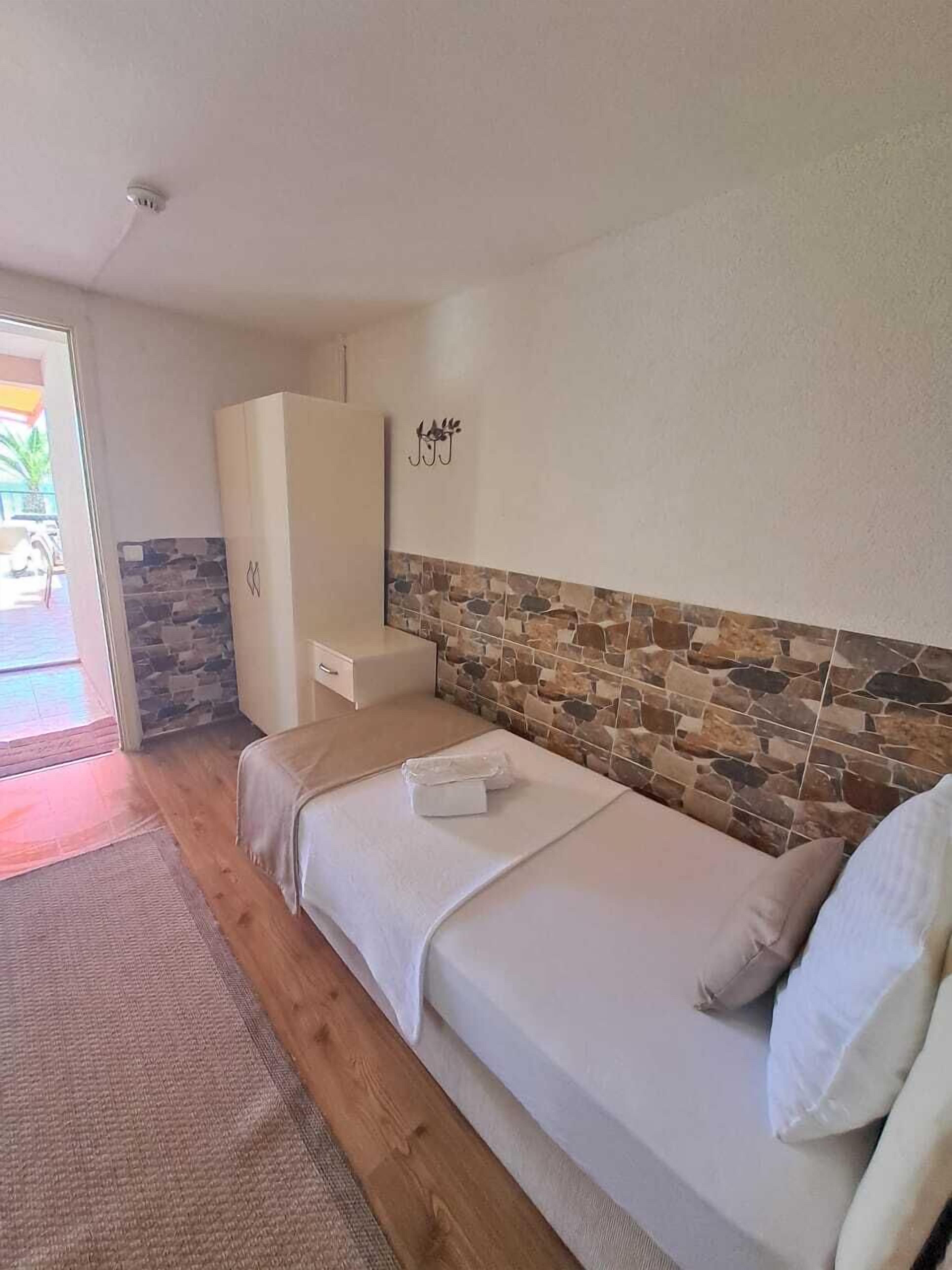 A bedroom with a wooden floor, a bed with white bedding, a stone accent wall, a white wardrobe, and a small table.
