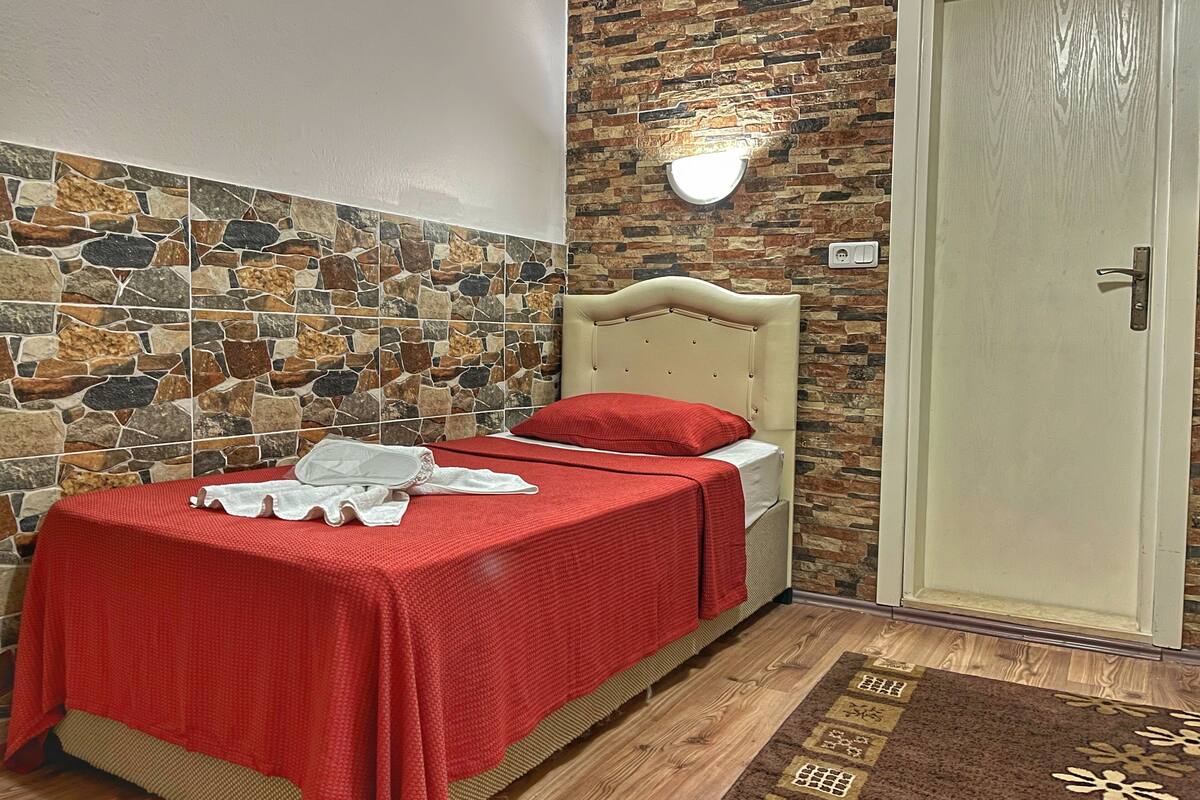 Une chambre avec un mur en pierre, un lit avec une literie rouge, une porte blanche et un petit tapis.