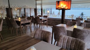 Un restaurante con mesas y sillas de madera, una gran pantalla de televisión mostrando un fuego, y vista al mar.