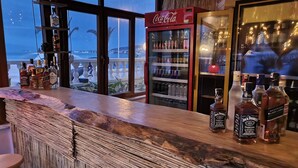 Un bar con mostrador de madera, estantes con botellas de licor, un refrigerador de Coca-Cola y vista a la playa.