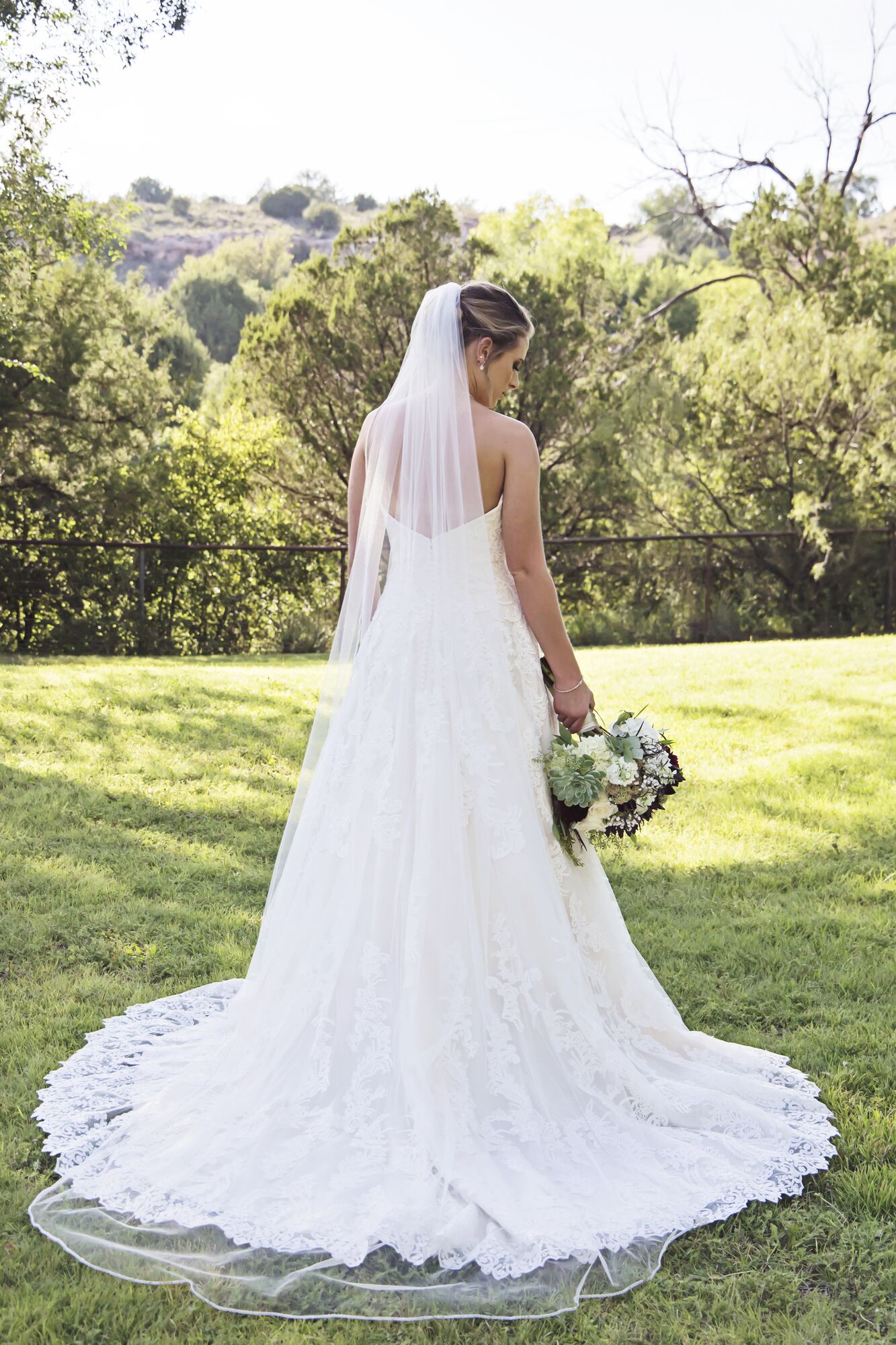 A bride in a white wedding dress with a long train, holding a bouquet, standing in a grassy outdoor area with trees in the background.