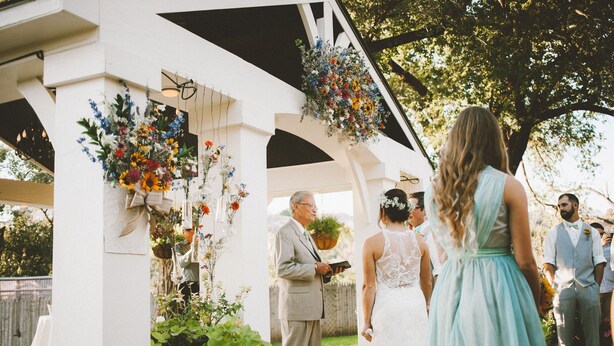 Una ceremonia de boda bajo un quiosco blanco, decorado con arreglos florales y follaje colgante.