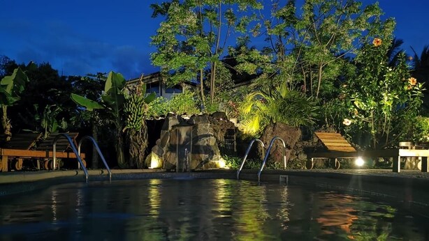 Una piscina con una cascada sobre rocas, rodeada de vegetación exuberante y una terraza de madera.