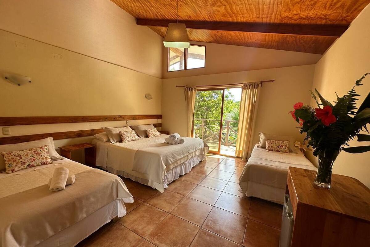 A hotel room with three single beds, a wooden ceiling, and a large window with curtains.