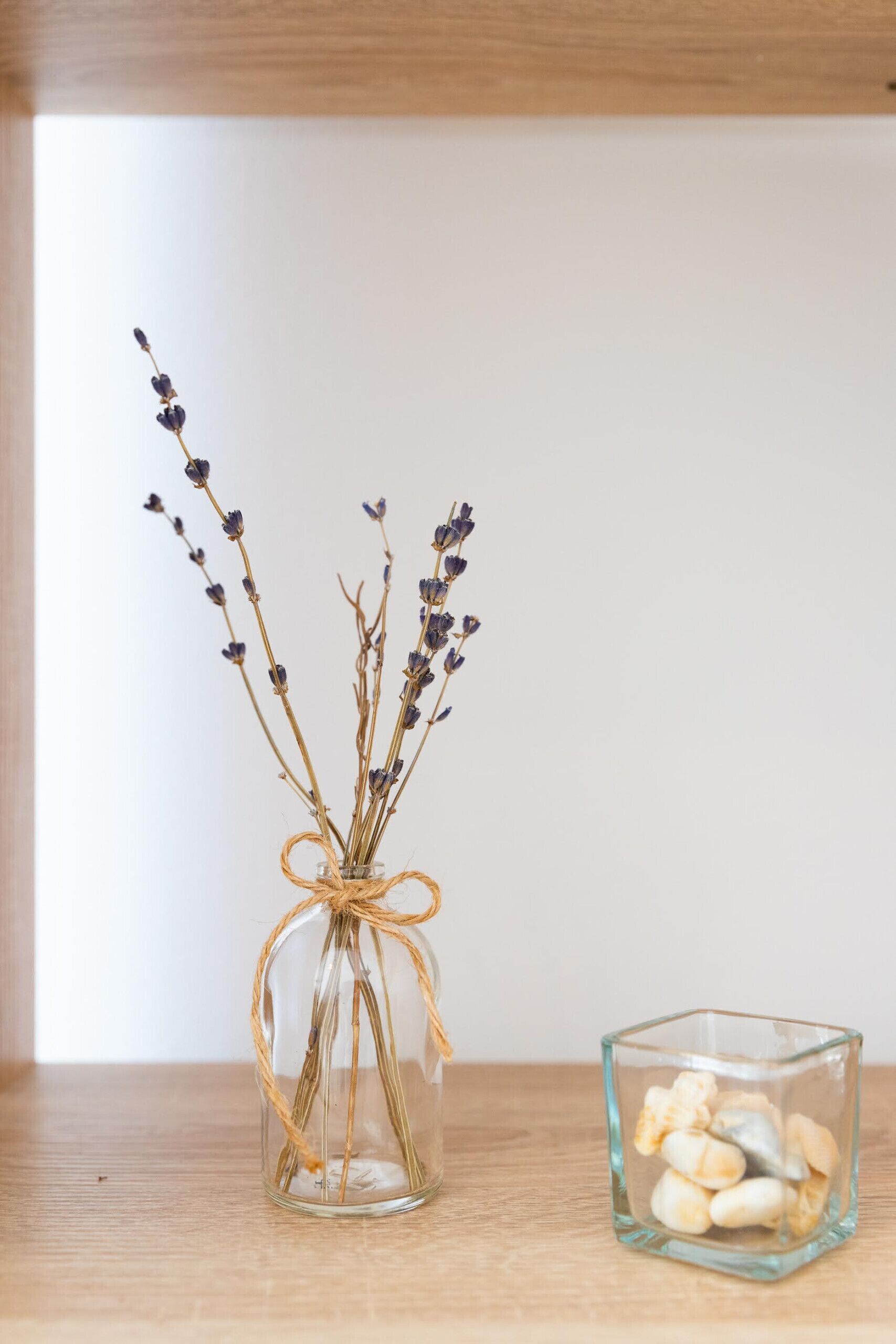 A clear glass vase with dried lavender stems tied with twine, placed on a wooden surface next to a glass cube containing small stones.