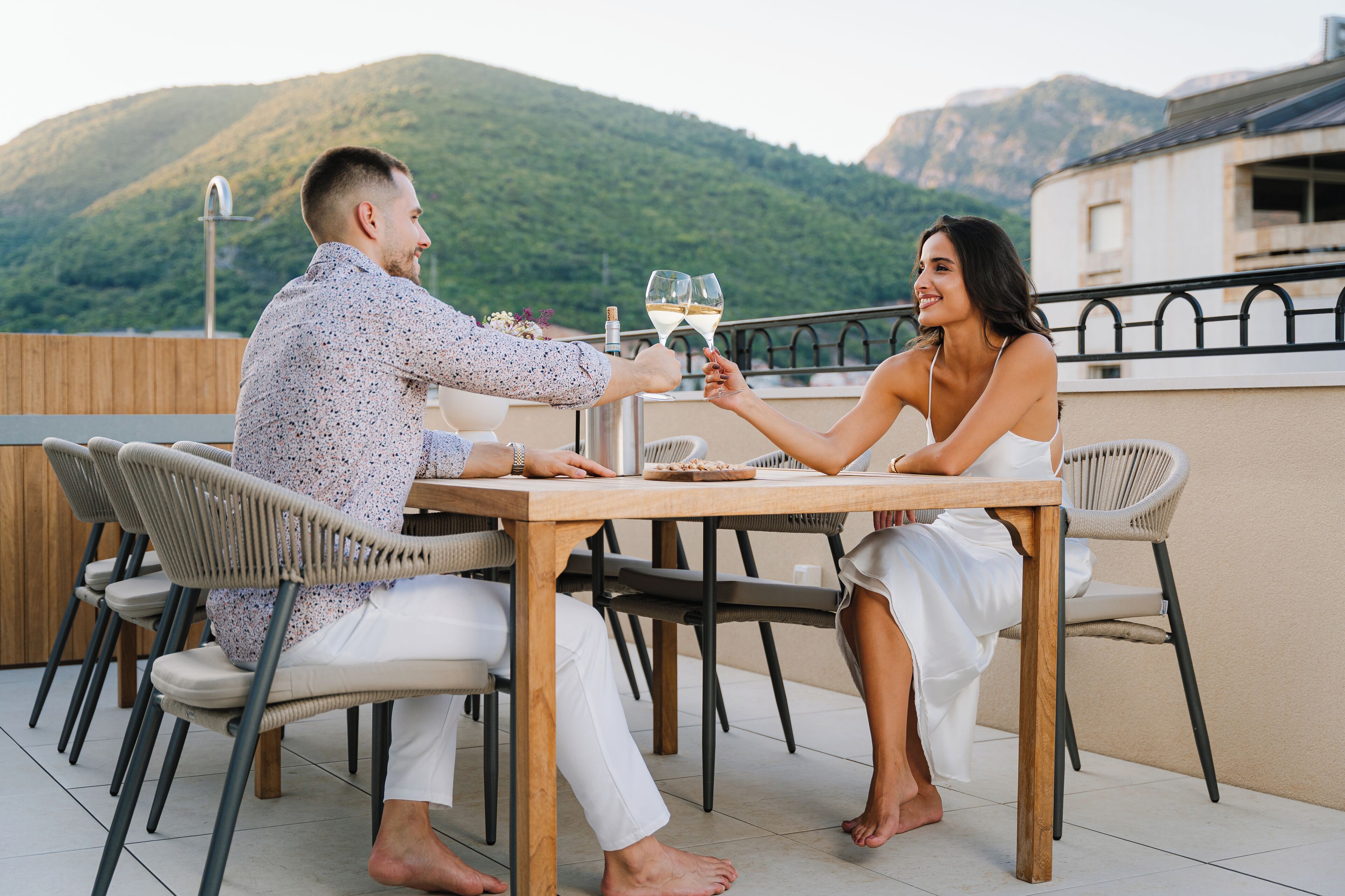 Un couple trinque à une table en bois, avec une chaîne de montagnes en arrière-plan.