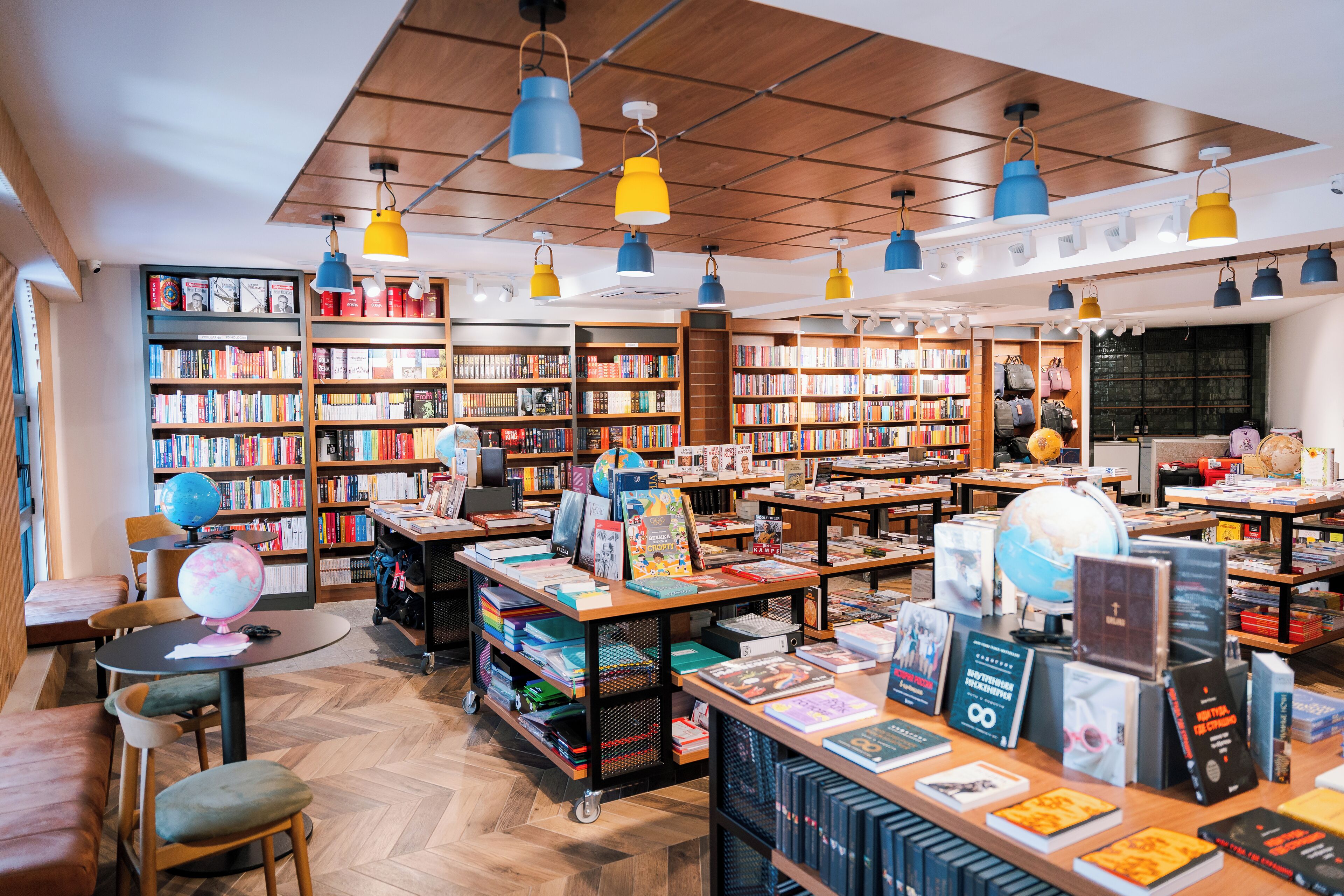 A bookstore with wooden shelves, tables, and chairs, and hanging blue and yellow lamps.
