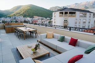Una terraza en la azotea con una mesa de madera, sillas, un sofá y vistas a un paisaje urbano y montañas.