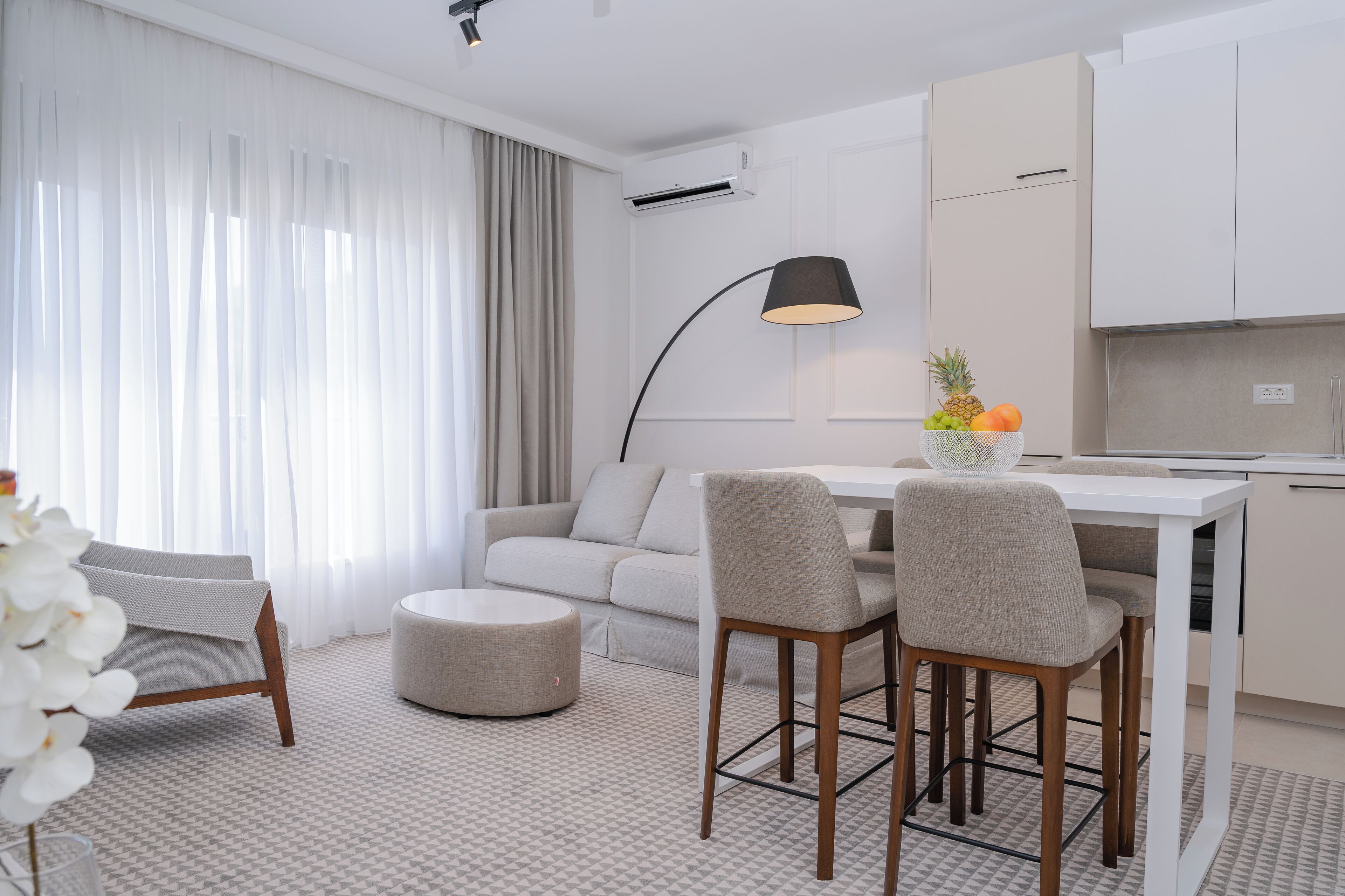 A modern, well-lit living area with a sofa, ottoman, and chairs, a dining table, and a kitchen area with a fruit bowl.