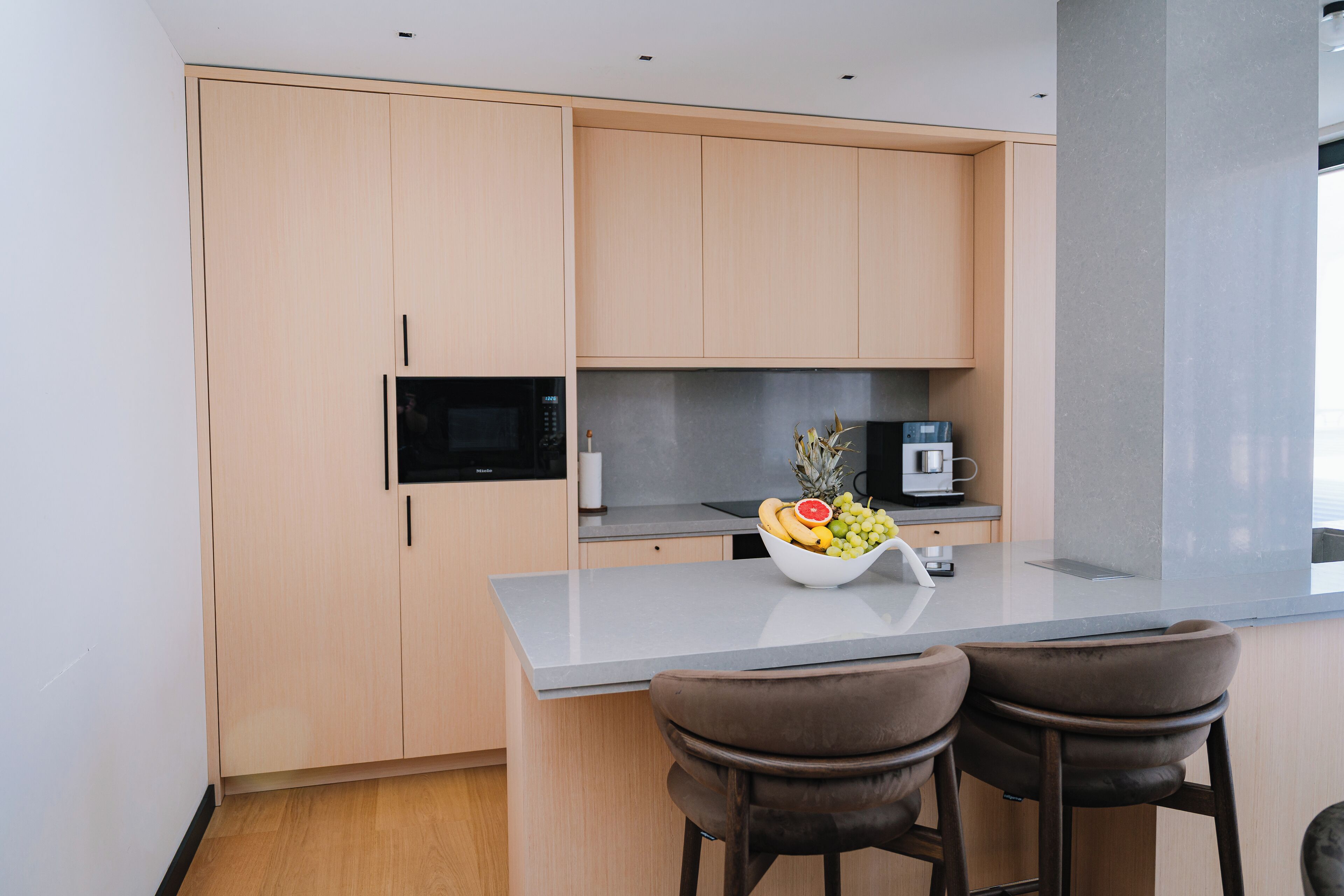 A modern kitchen with light wood cabinets, a grey countertop, and two bar stools.