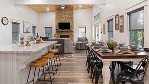 A spacious kitchen with a stone fireplace, wooden ceiling, and a large dining area with bar stools and a long table set for six. - Rustic Hillside Hideaway-2 BR+Bunk-Timber Creek (Broken Bow)
