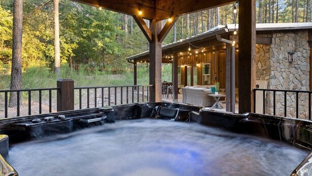 A hot tub area with a stone wall and wooden deck, surrounded by trees. - Rustic Hillside Hideaway-2 BR+Bunk-Timber Creek (Broken Bow)
