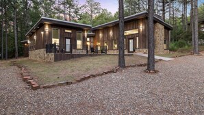 A modern cabin with a stone foundation and wooden exterior, surrounded by a gravel driveway and a wooden deck. - Rustic Hillside Hideaway-2 BR+Bunk-Timber Creek (Broken Bow)
